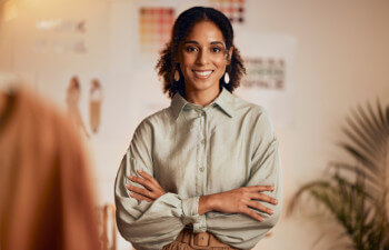 A woman stands indoors with arms crossed, smiling at the camera. She is wearing a light-colored blouse and earrings.