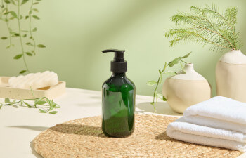 A green pump bottle on a woven mat next to folded white towels, ceramic vases, and green plants in a bright, minimalist setting.