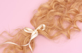 Blonde curly hair tied with a pale ribbon, placed on a pink background.