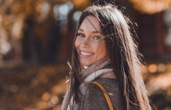 A woman with long brown hair smiles at the camera outdoors, wearing a coat and scarf on a sunny autumn day.