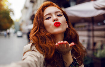 A woman with long red hair and red lipstick blows a kiss toward the camera while standing outdoors on a city street.