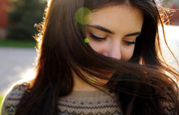 Young woman with long brown hair looks down, partially covering her face with her hair, outdoors in bright sunlight.