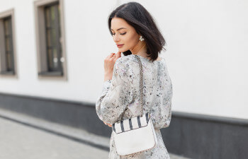 Woman with shoulder-length dark hair wearing a light floral dress and holding a white handbag with a chain strap, standing outdoors near a white wall.