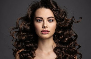 A woman with long, curly dark hair and neutral makeup is looking directly at the camera against a dark background.