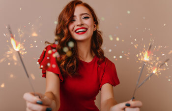 Woman in a red dress smiling and holding two lit sparklers against a plain beige background.