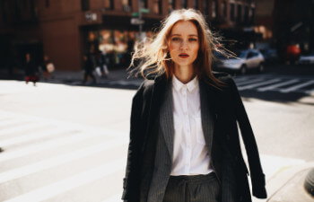 A woman in a white shirt and dark blazer stands at a crosswalk in a New York City.