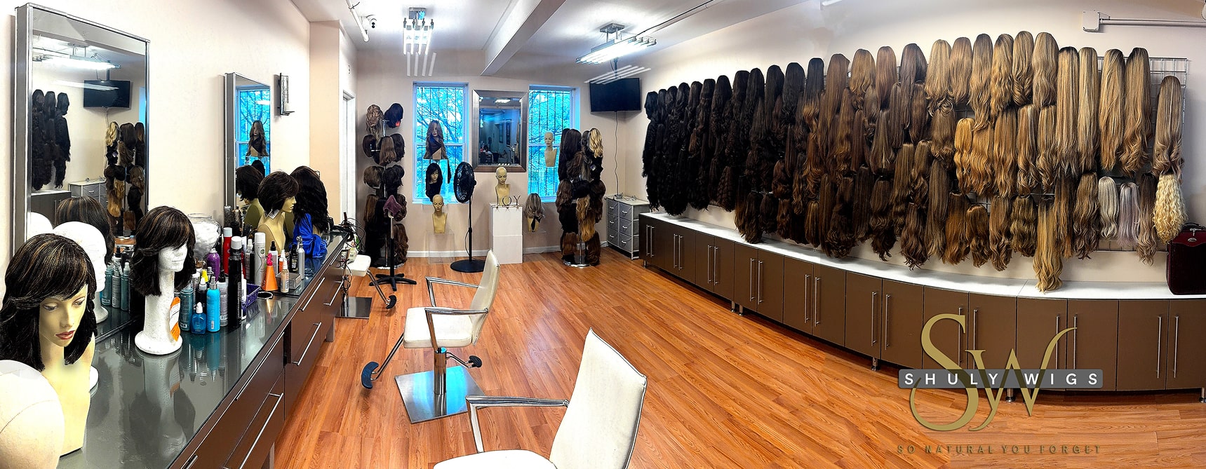 A wig shop interior with rows of wigs on the wall, styling chairs, mirrors, mannequin heads with wigs, and various hair products on the counter.