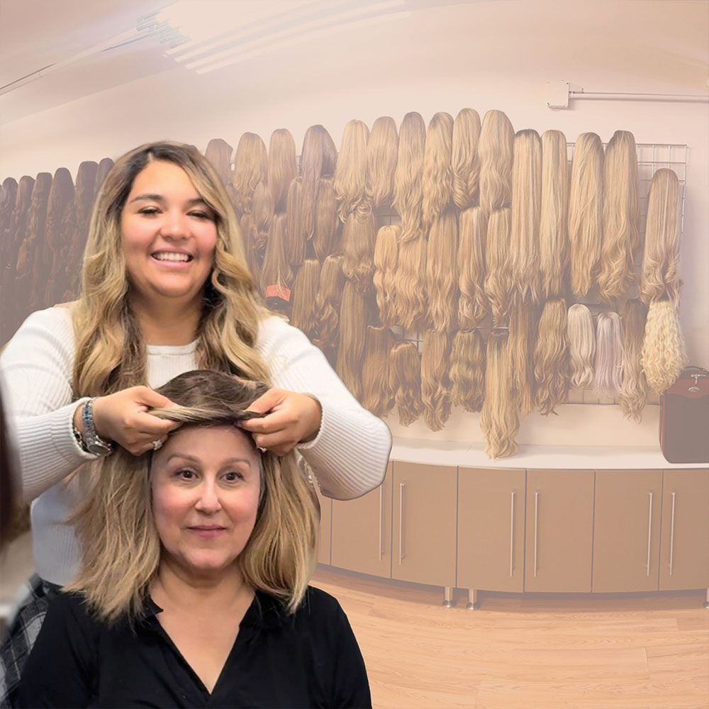 A smiling woman fits a wig on another seated woman in a room with many wigs displayed on the wall behind them.