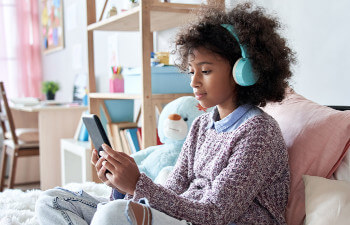 A young girl wearing blue headphones sits on a bed and looks at a tablet.