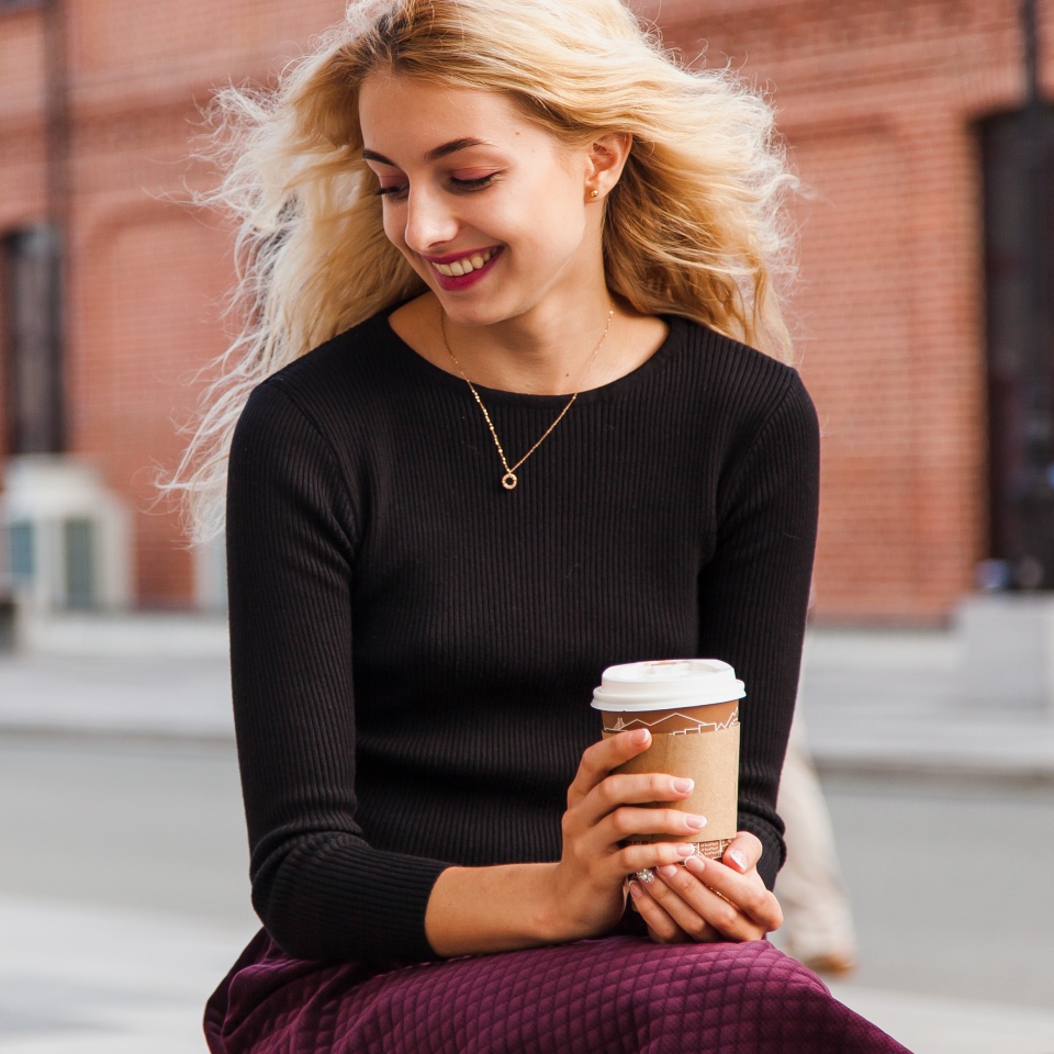Attractive pretty girl in her early 20s looks down waiting for meeting with cup of coffee to go wearing modest casual clothes. Smiley young woman having good mood
