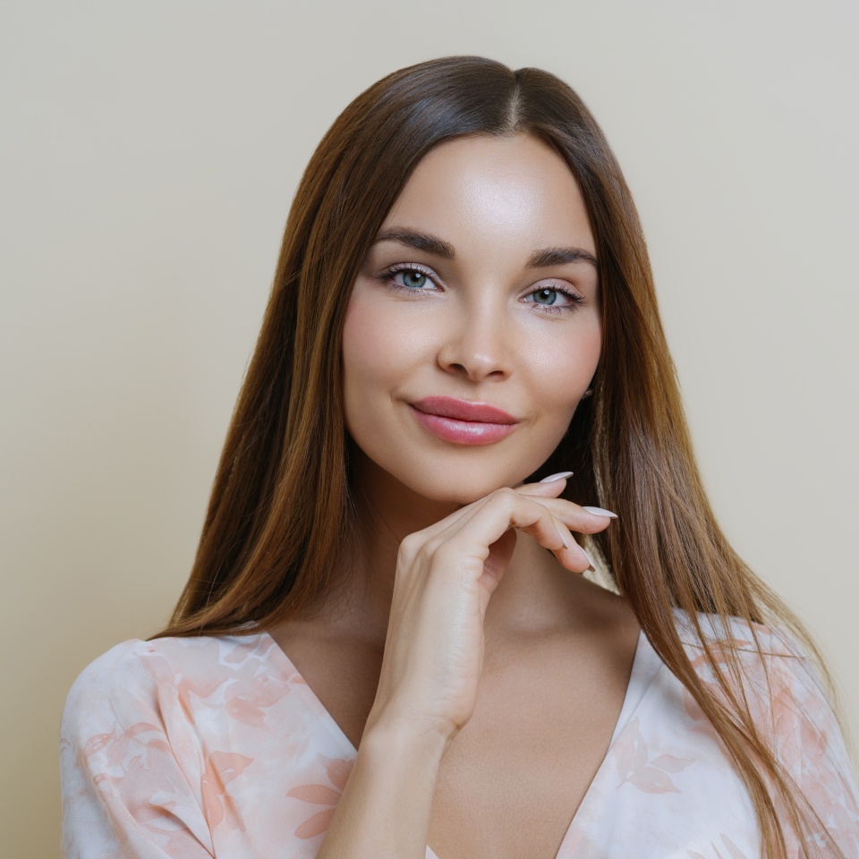 Natural beauty and people concept. Portrait of beautiful dark haired young European woman touches jawline gently, dressed in stylish dress, isolated over beige background, dreams about perfect day