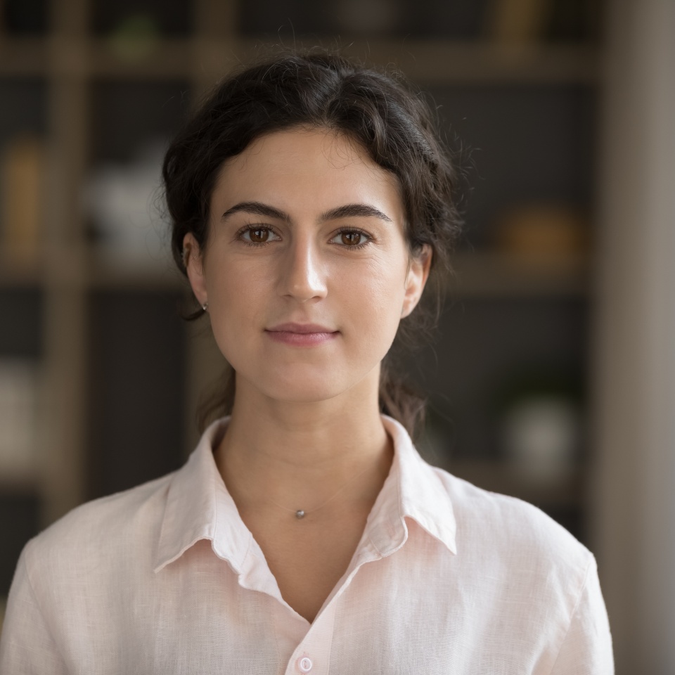 Head shot serious good-looking young 20s woman in casual elegant shirt standing alone in cozy living room staring at camera feels confident pose indoor. Housemaid or real-estate agent portrait concept