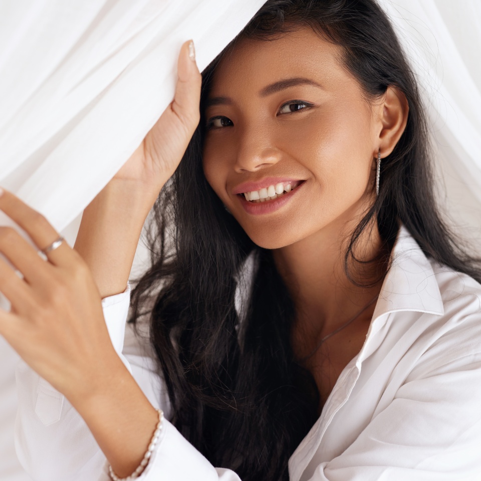 Happy Woman With Beautiful Face Smiling, Lying On Bed. Portrait Of Beautiful Young Female With Healthy Hair, Fresh Soft Facial Skin And Perfect White Smile On White Bedding. High Resolution.