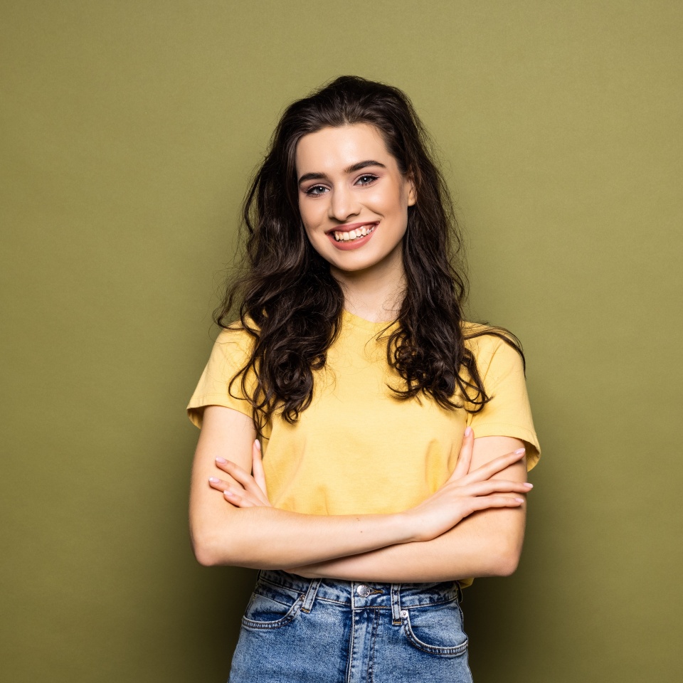 Young smiling attractive girl is posing in close pose with arms crossing on chest on khaki background looking at camera. Shy student, modest woman.