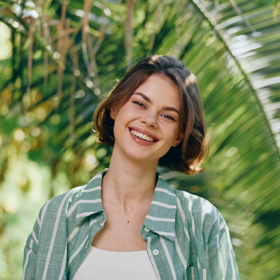 Smiling woman outdoors in natural light, wearing a striped green jacket over a white top, framed by lush tropical greenery and sunlit leaves.