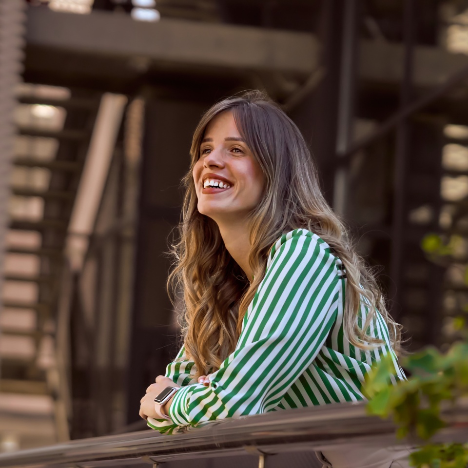 Smiling Woman Leaning on Railing in Green Striped Shirt Outdoors contemplating. Portrait of a confident young businesswoman in front of the modern office building.