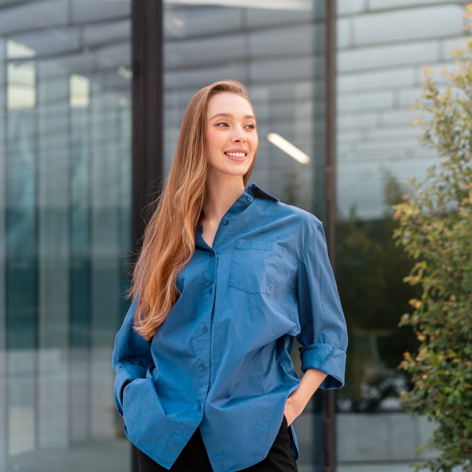 Smiling woman in blue shirt standing with hands in pockets outside modern office, looking away with confidence and calm, reflecting professionalism and smart casual style