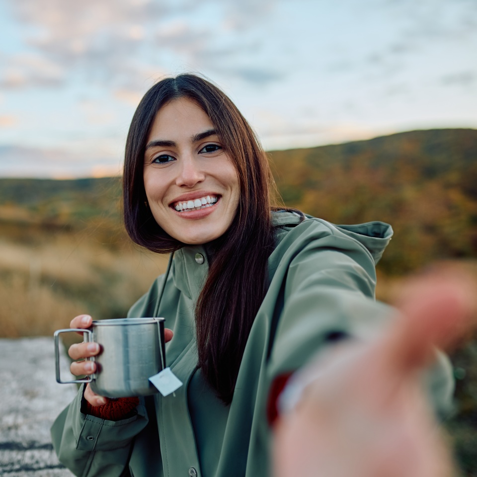 Young woman holding metal cup with tea, taking a selfie while smiling happily, enjoying nature during an outdoor activity