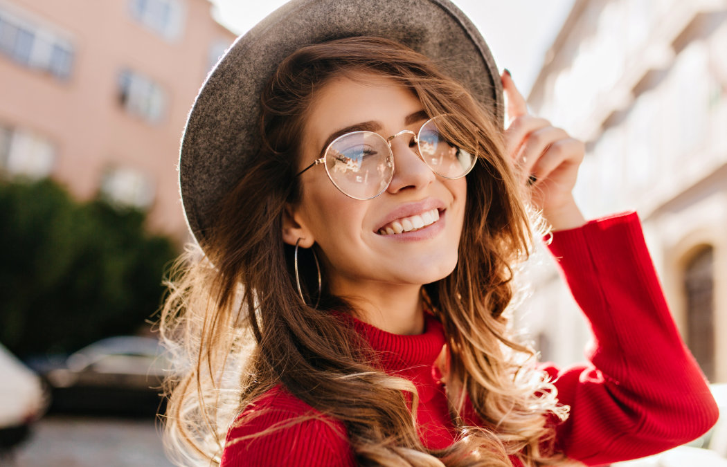 Woman in a red sweater smiles while touching her hat. She is wearing round glasses and hoop earrings
