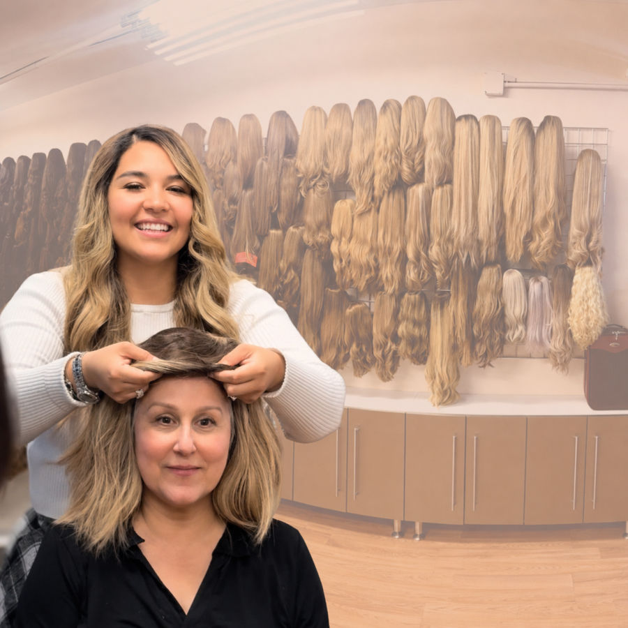 A smiling woman fits a wig on another seated woman in a room with many wigs displayed on the wall behind them.